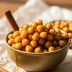 Golden honey roasted chickpeas in a bowl with honey drizzle and cinnamon stick on parchment paper.